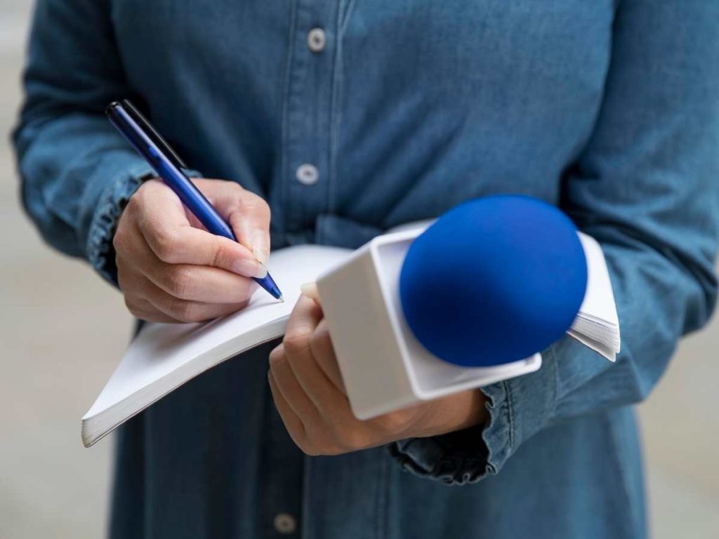Pessoa escrevendo em um caderno e segurando um microfone azul, representando a seleção de tema para uma tese de jornalismo.