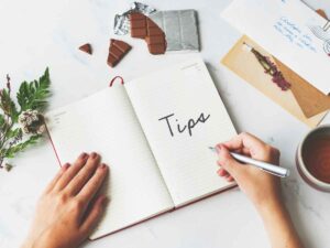 Pessoa escrevendo a palavra dicas em caderno aberto sobre mesa com chá, chocolate e cartas, simbolizando conselhos para projetos de mestrado.