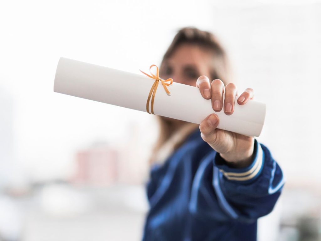 Menina ao fundo pegando e exibindo seu certificado de aprovação fechado