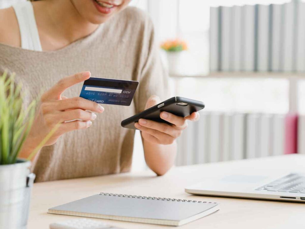 Pessoa segurando cartão de crédito e smartphone ao lado de laptop, representando o pagamento online de serviços acadêmicos.