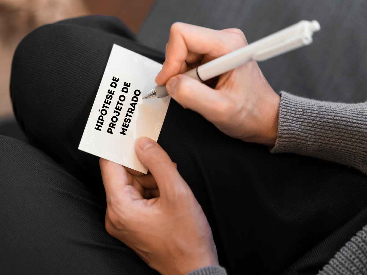 Pessoa escrevendo na folha com a frase "HIPÓTESE DE PROJETO DE MESTRADO" sendo anotada.