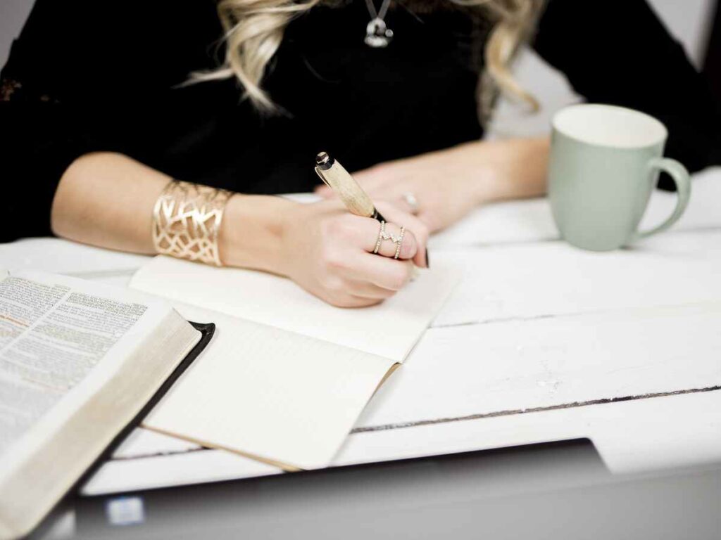 Mujer escribiendo en una libreta mientras toma una taza de café, con un ordenador portátil en la mesa