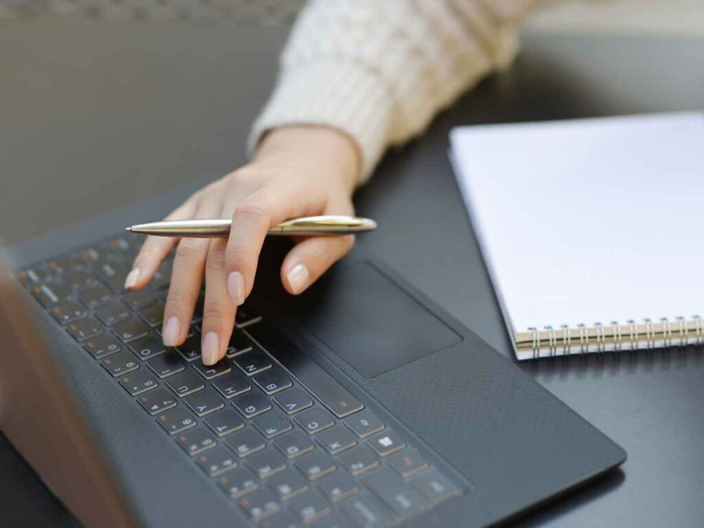 Pessoa trabalhando no teclado de um laptop com uma caneta na mão