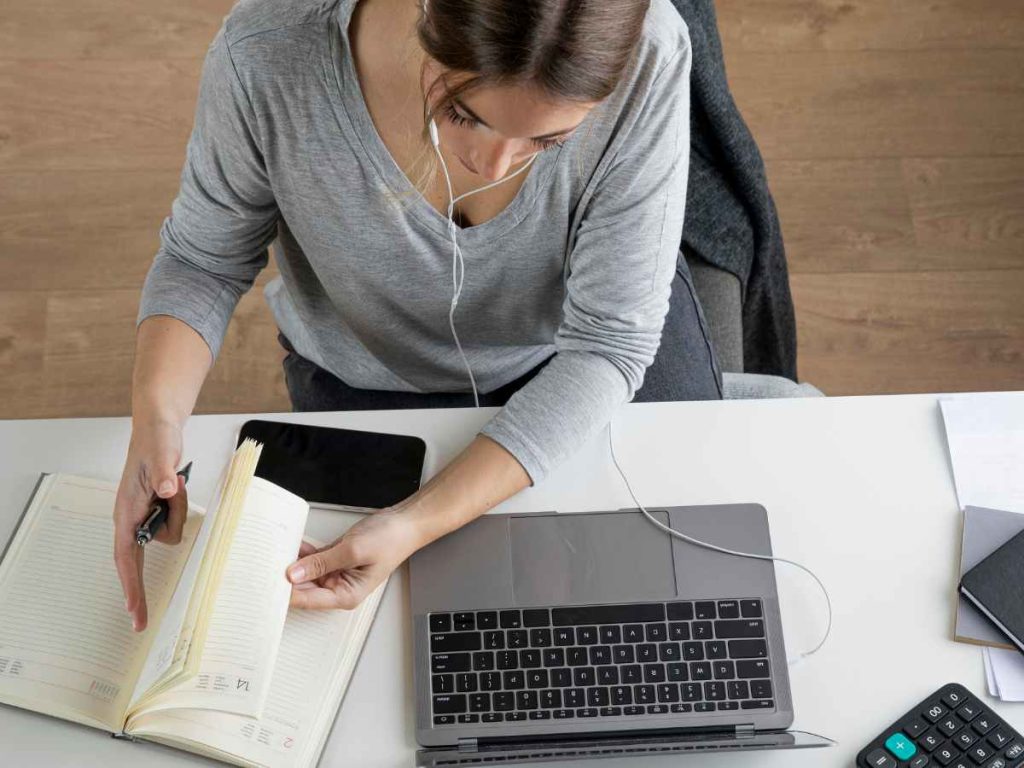 Estudante escrevendo em um caderno de forma detalhada, com computador ao fundo para elaboração de projeto de mestrado.